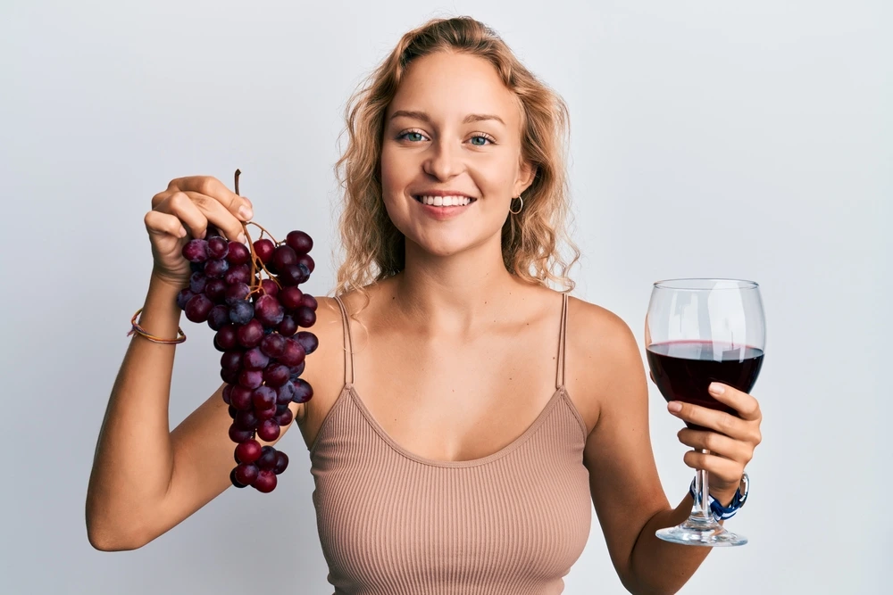 Cute girl holding red wine in a glass and a cluster of red grapes while smiling